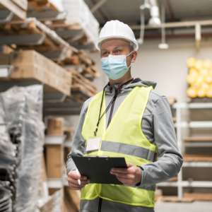 Warehouse employee wearing a covid mask, hard hat and high visibility vest. He is holding a clipboard and looking firmly ahead into the warehouse