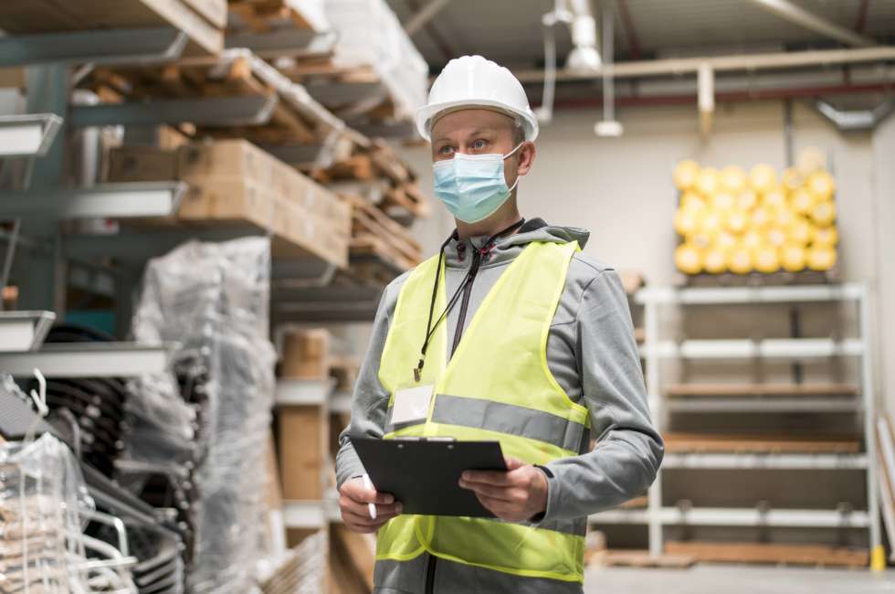 Warehouse employee wearing a covid mask, hard hat and high visibility vest. He is holding a clipboard and looking firmly ahead into the warehouse