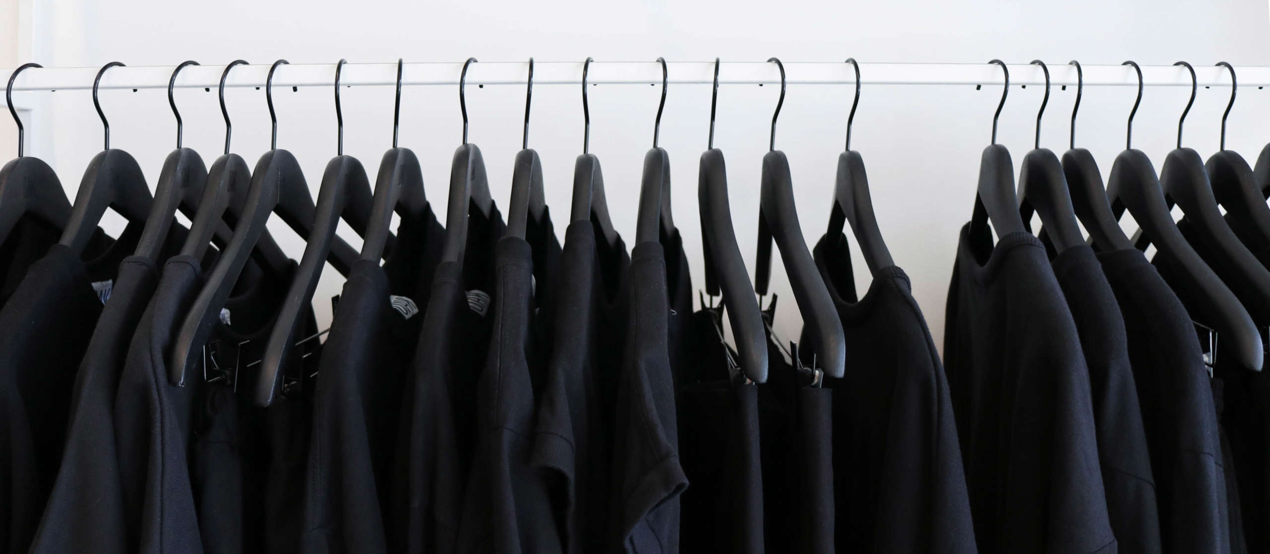 Black clothing that is hung up on matching black hangers, the clothing is hanging off of a white rail contrasting a white wall