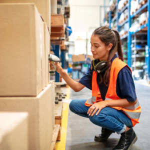 Warehouse worker crouching down and checking boxes on shelves with scanner. Female worker scanning boxes in rack.